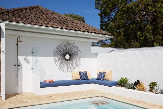 Bright outdoor courtyard with a small swimming pool beside a white rendered building. The space features a built-in blue L-shaped bench with cushions, a wall-mounted sunburst decoration, a frameless glass shower screen, pale stone paving, white boundary walls, and a brown curved terracotta tile roof. Established trees and sculptural planting surround the courtyard.