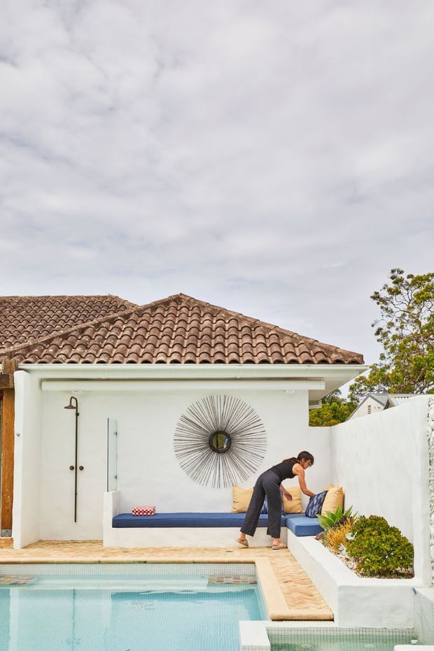 Exterior courtyard beside a swimming pool, featuring a white rendered cabana or poolhouse with a terracotta barrel-tile roof, built-in blue-cushioned seating, outdoor shower, decorative sunburst mirror, white masonry walls, and landscaped planters. A person is arranging cushions on the seating.