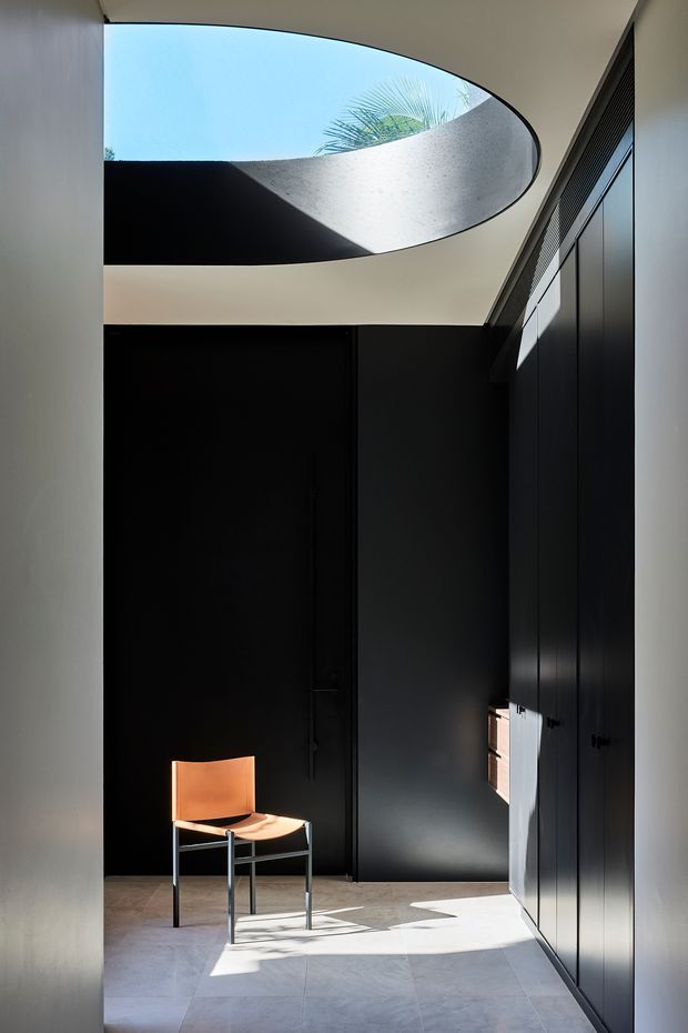 A minimalist interior passage or entry space with dark full-height built-in cabinetry and a black door, contrasted by pale stone or concrete floor and light walls. A sculptural oval skylight opens to blue sky and palm foliage, casting strong sunlight and shadows across the floor. A single orange-brown chair provides the main accent.