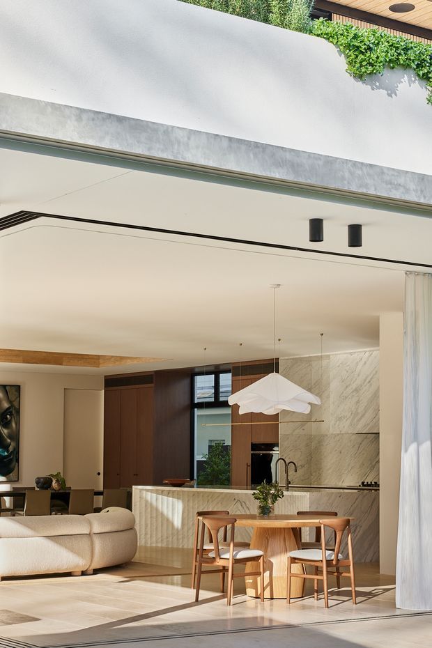 Bright contemporary open-plan interior viewed from a covered outdoor edge, combining a living room, dining area and kitchen. The space features a cream sectional sofa, warm timber cabinetry and dining furniture, a pale stone kitchen island and full-height marble-look wall, large glazed openings, concealed linear lighting, and a sculptural white pendant above the dining table. A planted upper terrace and timber-lined soffit are visible above the opening.