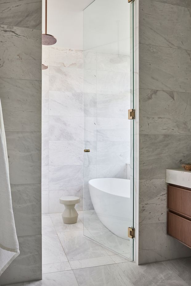 A refined contemporary bathroom featuring a glass-enclosed shower and freestanding white bathtub. Large-format pale grey and white veined stone tiles cover the walls and floor, complemented by a floating timber vanity with a light stone benchtop and warm brass hardware. A small sculptural stool and pendant light add subtle decorative detail.