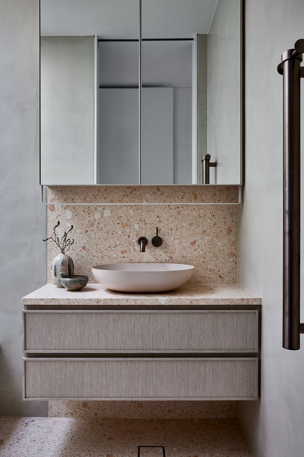 A compact, refined bathroom featuring a floating double-drawer vanity in a textured taupe-grey finish, a pale terrazzo countertop and matching full-height splashback, a white vessel basin, dark bronze wall-mounted tapware, and mirrored wall cabinets. A dark timber-framed doorway or screen is visible at right.