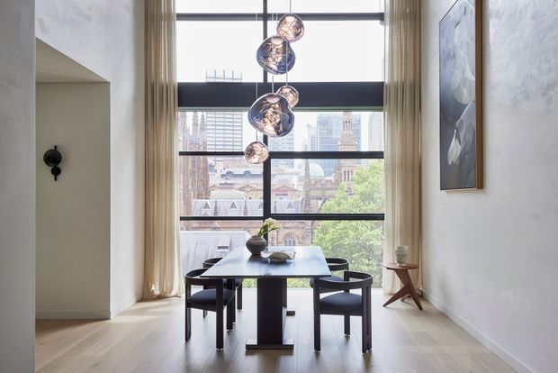 A refined dining area with a dark rectangular dining table and four black upholstered chairs positioned in front of floor-to-ceiling glazing. Tall beige curtains frame a broad city view, while sculptural pendant lights hang above the table. Pale timber flooring, white textured walls, minimalist artwork, and a small timber side table create a bright, elegant interior.