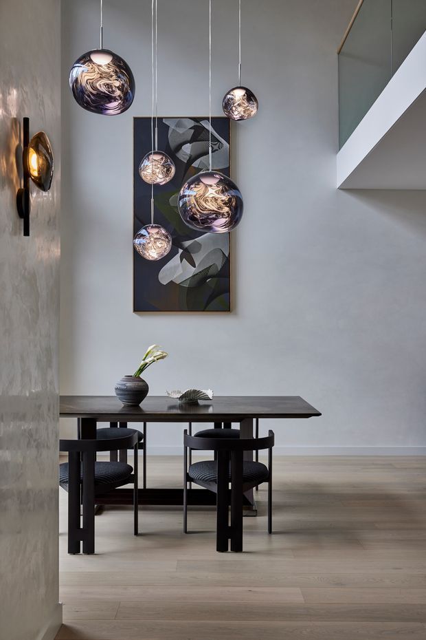 A refined contemporary dining area viewed from an adjoining space, featuring a dark rectangular dining table with black chairs, pale timber flooring, smooth light-grey walls, and a dramatic cluster of suspended spherical pendant lights. Abstract artwork hangs behind the table, while a sculptural wall light and minimalist decor add visual interest. A partial upper-level opening or stair void is visible at right.