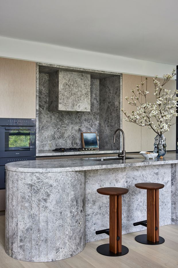 A refined contemporary kitchen featuring a large curved island clad in richly veined grey stone, with matching stone splashback and integrated rangehood surround. Light timber cabinetry spans the walls, while dark built-in appliances, a sculptural black tap, and two timber-and-black bar stools add contrast. A decorative flowering branch in a vase provides a soft organic accent. The space is bright, polished, and luxurious.