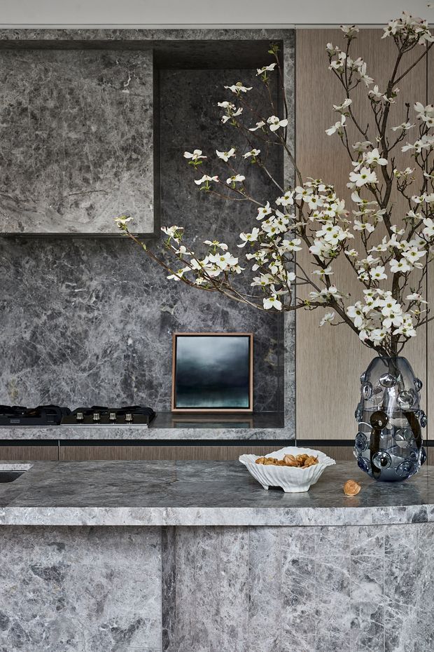 A refined contemporary kitchen featuring extensive grey veined natural-stone wall cladding and benchtops, dark cabinetry, and a textured vertical timber-toned panel. A black cooktop is integrated into the rear counter, while the foreground island has a thick stone waterfall-style edge. Decorative branches in a sculptural glass vase and a small framed artwork add contrast to the moody, high-end composition.