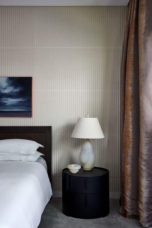 A sophisticated bedroom vignette featuring a partially visible bed with crisp white bedding and a dark timber headboard, a black round bedside table, a sculptural white ceramic lamp, and a small decorative bowl. The wall is clad in pale vertically ribbed fluted panels, complemented by full-height dark brown curtains and grey carpet. A moody framed artwork adds contrast. The room has a refined, contemporary aesthetic with layered neutral tones and strong textural detail.