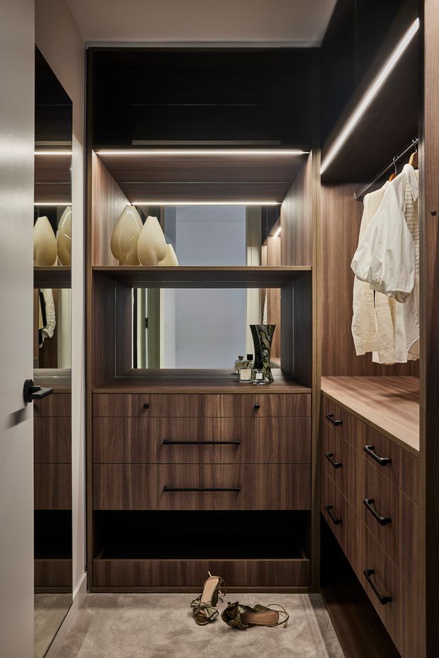 A compact, highly organised walk-in wardrobe with dark walnut-toned timber cabinetry, open hanging rails, drawers, shelving, integrated warm LED strip lighting, and a central mirrored dressing niche. Neutral carpeting and a few clothing items and shoes are visible.
