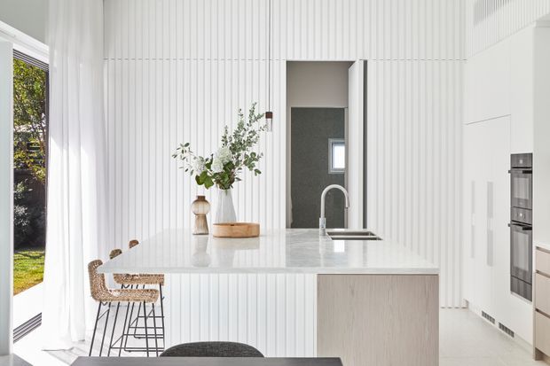 Bright contemporary kitchen with a large pale stone island, integrated sink and curved faucet, light timber cabinetry, white vertical fluted wall panelling, and built-in stainless-steel appliances. A small dining or breakfast area with stools is visible beside floor-to-ceiling glazing and sheer curtains.