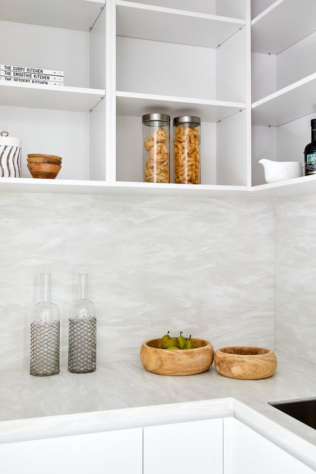 Bright, minimalist kitchen corner with open white shelving, a pale marbled splashback and matching benchtop, and white lower cabinetry. The shelves display pasta jars, bowls, bottles and small decorative objects, while wooden bowls and glass bottles sit on the benchtop.