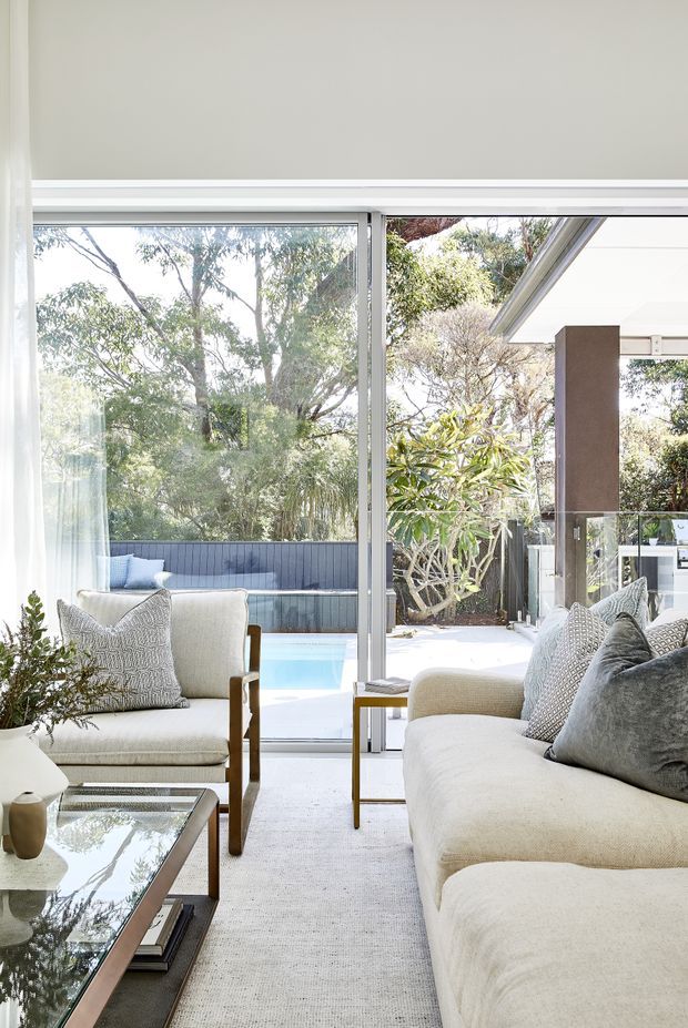 A bright, refined living room with a cream sofa, matching upholstered armchair, patterned grey cushions, glass-topped coffee table, and light carpet. Full-height sliding glass doors open directly to an outdoor terrace with a swimming pool and landscaped garden. The room has a neutral palette, abundant natural light, and a strong indoor-outdoor connection.