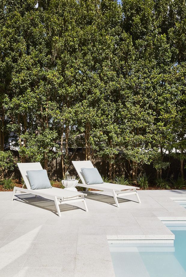 A bright outdoor pool area with two white reclining loungers and pale cushions positioned on a large light-coloured tiled deck beside a swimming pool. Dense mature greenery forms a private garden backdrop, with subtle planting along the edge.