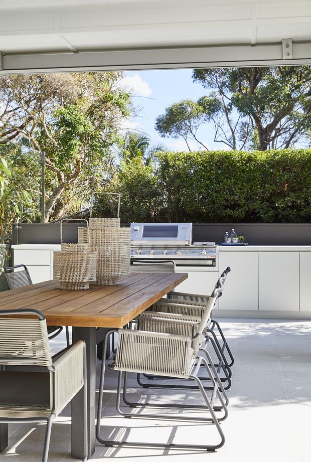 A bright contemporary outdoor entertaining area beneath a raised garage-style door, featuring a built-in stainless steel barbecue set into white cabinetry, a long timber dining table with woven outdoor chairs, pale tiled flooring, and a landscaped hedge and trees beyond.