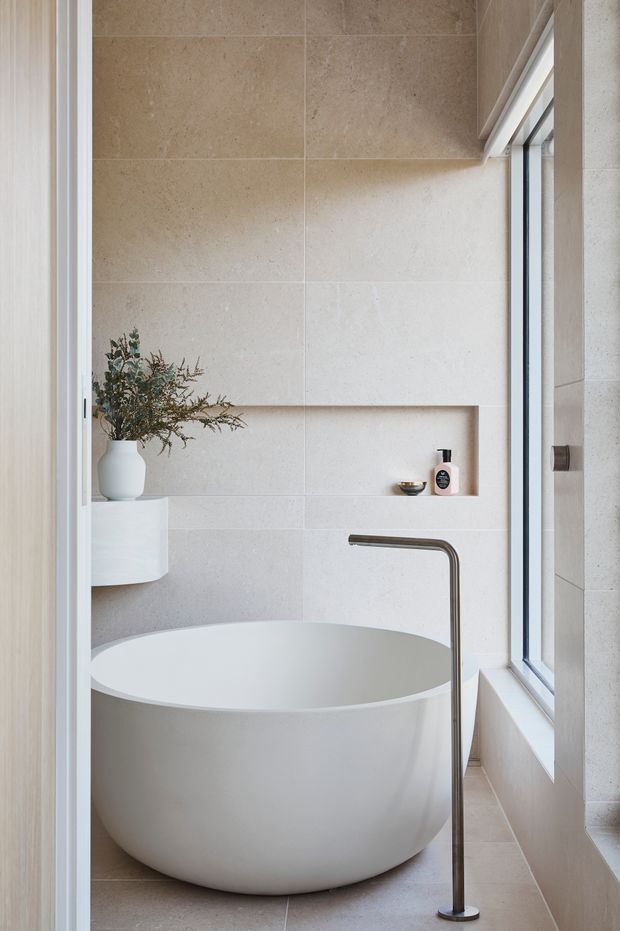 A serene, minimalist bathroom featuring a freestanding oval white bathtub beneath a large window. The room is clad in large warm beige and off-white stone-look tiles, with a slim dark metal floor-mounted bath filler, recessed wall niche, and a simple white vessel basin partially visible beside a vase of foliage.