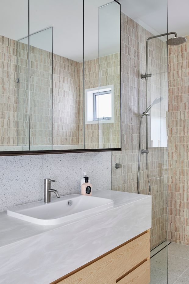 A bright, contemporary bathroom featuring a large frameless glass shower enclosure, vertically patterned beige and blush ceramic wall tiles, a white integrated basin set into a pale stone-look vanity, timber cabinetry, a wall-mounted mirror, and brushed metal shower and tapware.