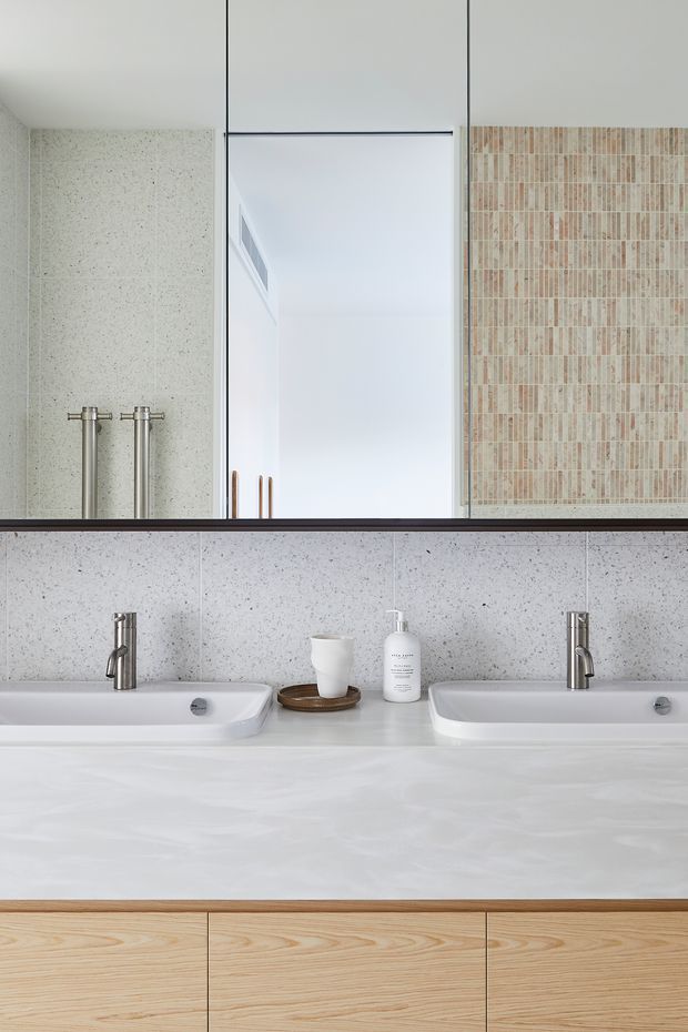 Contemporary double vanity bathroom with two white integrated basins, slim chrome tapware, a broad white stone benchtop, light timber cabinetry, speckled pale wall surfaces, and a large frameless mirror. A warm-toned vertical mosaic tile feature is reflected in the mirror, adding subtle texture and contrast.