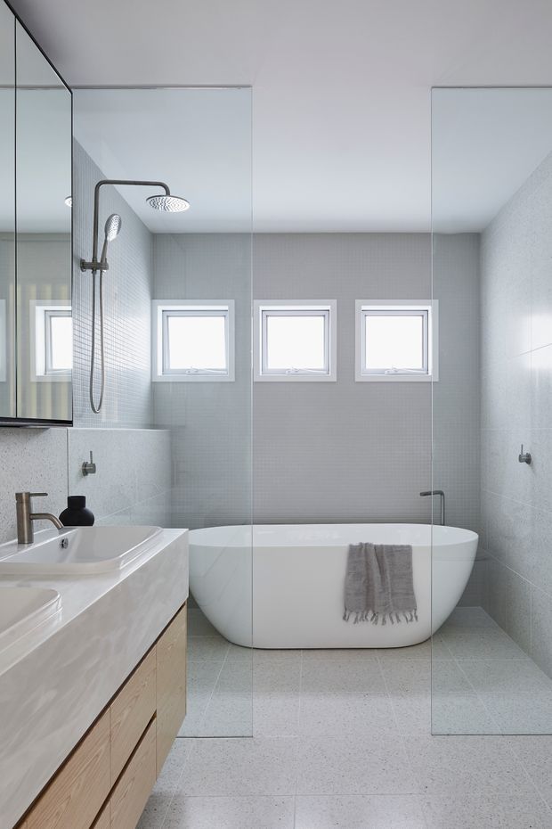 Bright contemporary bathroom with a freestanding white bathtub, wall-mounted shower, large frameless glass shower enclosure, double vanity with pale timber cabinetry, pale grey tiled walls and floor, and three high-set windows. The room has a clean, minimal aesthetic with brushed metal fixtures and soft grey textiles.