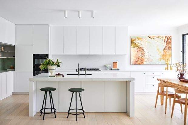 A bright, refined open-plan kitchen and dining area with extensive white flat-panel cabinetry, a large central island with two dark stools, integrated appliances, pale timber flooring, and a light timber dining table. An abstract artwork and indoor plants add warmth to the predominantly white interior.