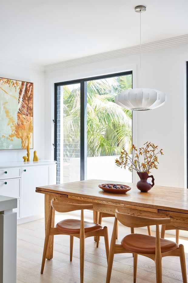 Bright, refined dining area with a large solid timber dining table and matching sculptural wooden chairs. The room features white walls and cabinetry, pale timber flooring, dark-framed floor-to-ceiling glazing, decorative ceiling moulding, a contemporary pendant light, and a large abstract artwork. Palm foliage is visible outside the windows.