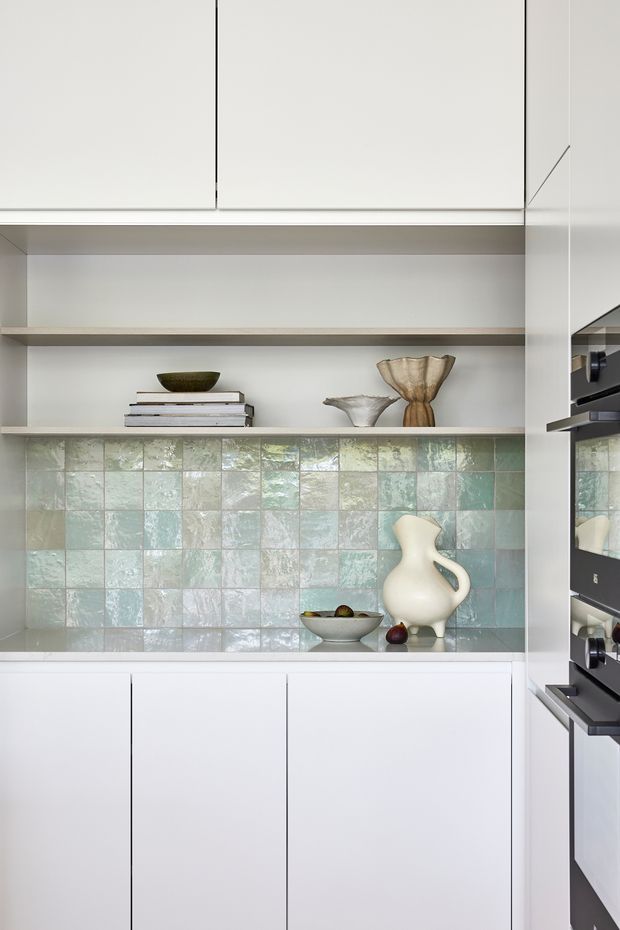 A refined contemporary kitchen with flat-front white cabinetry, open timber-toned display shelving, and a pale green and blue handmade-look tiled splashback. Sculptural ceramics, stacked books, and a fruit bowl provide restrained styling. Integrated black appliances are visible at the right edge, with a mirrored panel reflecting the kitchen.
