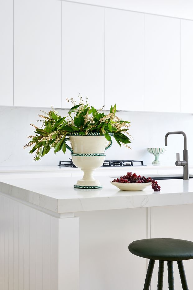 Bright, predominantly white kitchen interior with a large island, white cabinetry, a gas cooktop, and a dark metal gooseneck-style tap. The island has a white stone benchtop and vertical panel detailing, with a dark green upholstered stool. A decorative urn filled with greenery and flowers and a bowl of grapes sit on the island, adding a traditional decorative note to the otherwise clean, refined space.