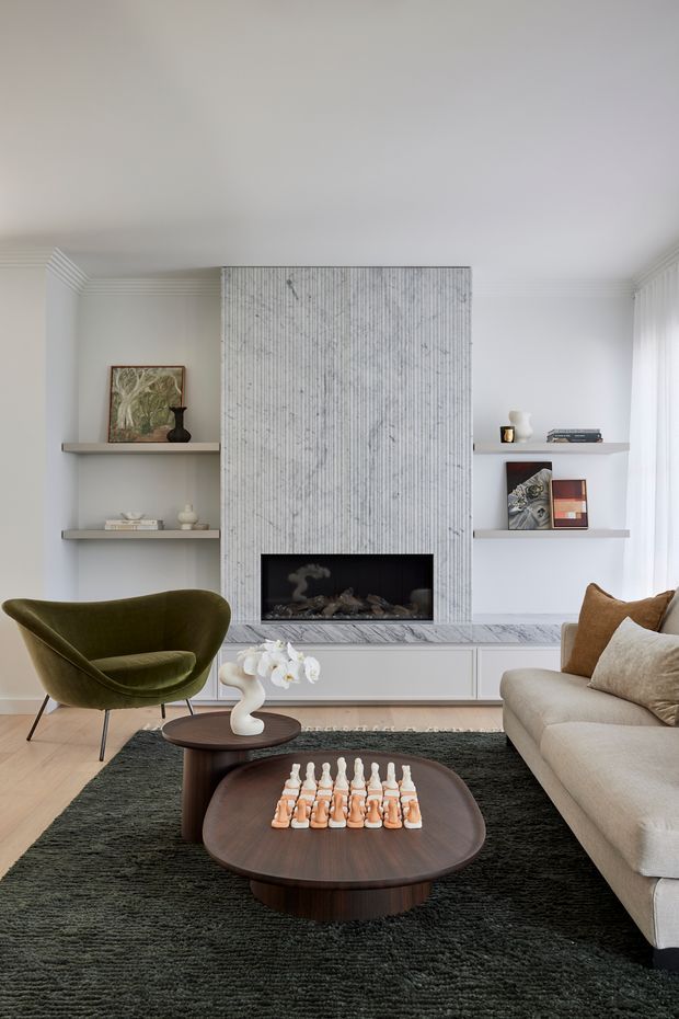 A refined living room with a contemporary fireplace feature wall clad in vertically textured light stone, flanked by minimalist open display shelves. The room includes a curved olive-green lounge chair, a large neutral upholstered sofa, dark sculptural timber coffee tables, a deep charcoal textured rug, pale timber flooring, and restrained decorative objects. Soft daylight enters through sheer curtains, creating a calm, sophisticated atmosphere.
