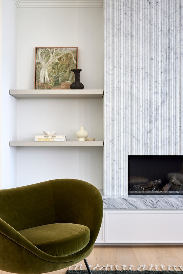 Refined living room vignette featuring a sculptural olive-green velvet lounge chair, built-in open display shelving, and a contemporary fireplace surround clad in vertically veined pale marble. The shelves hold artwork, books, ceramics, and a dark sculptural vase. Light timber flooring, white wall panelling, and a muted neutral palette create an elegant, softly layered interior.