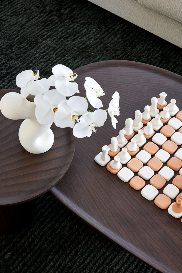 Close-up styled living room vignette showing overlapping dark timber coffee tables on a black textured rug, a white orchid arrangement in a rounded vase, and a contemporary decorative chess set in white and terracotta tones. A light upholstered sofa is partially visible at the top edge.