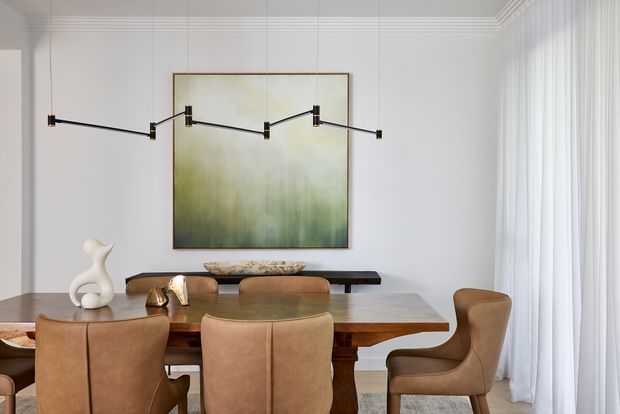 A refined dining room with a large solid timber dining table surrounded by tan leather upholstered chairs. A long, articulated black wall light is mounted over a softly abstract green and cream artwork, with sheer floor-to-ceiling curtains at the right and white vertically panelled walls with decorative cornice detailing.