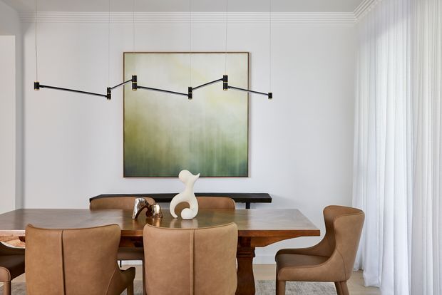 Refined dining room with a substantial warm timber dining table, tan leather upholstered chairs, a slim black console, and a large soft green abstract artwork. White walls, decorative ceiling moulding, sheer full-height curtains, and a sculptural linear black pendant create a bright, contemporary setting.