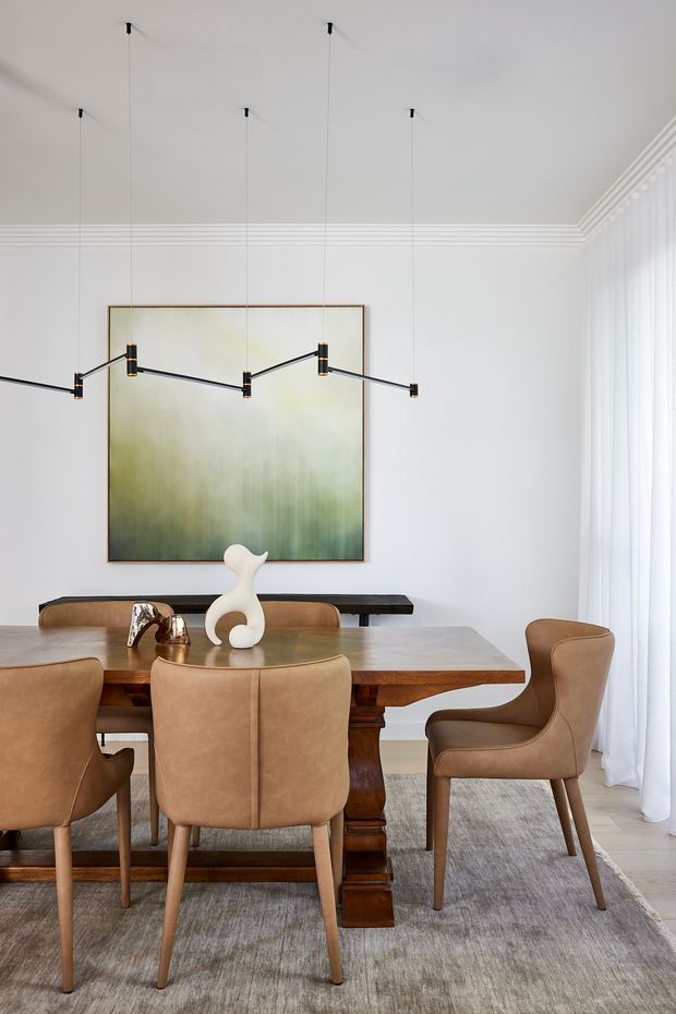 A refined dining room with a substantial dark timber dining table, upholstered tan leather dining chairs, a contemporary linear pendant light, and a large abstract green-toned artwork. White walls, ornate ceiling cornices, sheer floor-to-ceiling curtains, and a neutral rug create a bright, polished setting.