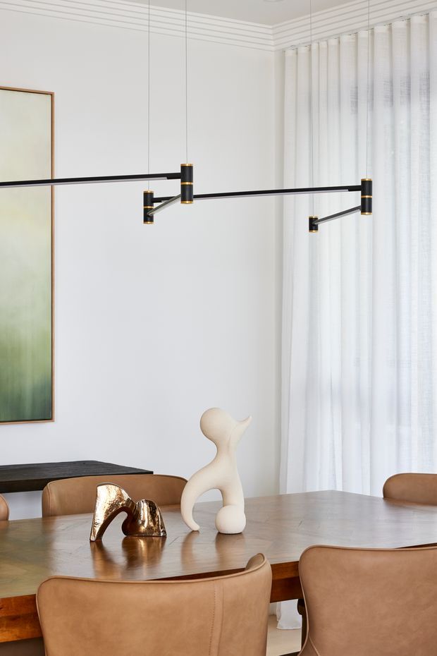 A refined dining area with a large timber dining table and upholstered tan leather dining chairs. A sculptural black linear pendant light with brass accents hangs overhead, while sheer white curtains cover a tall window. White walls feature subtle horizontal decorative moulding, with abstract artwork and sculptural objects adding visual interest.