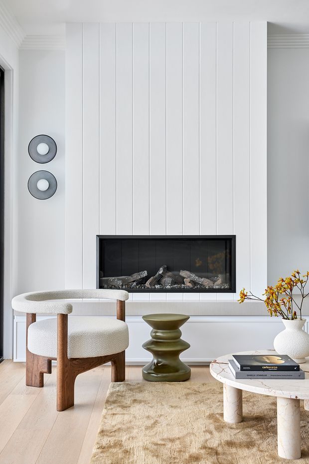 A refined living room with a white vertical-panelled fireplace wall and long black-framed built-in fireplace. The room includes a boucle upholstered timber armchair, sculptural olive-green side table, pale stone coffee table, neutral textured rug, and a white ceramic vase with branches. The palette is soft white, light timber, beige, olive green, and black accents.