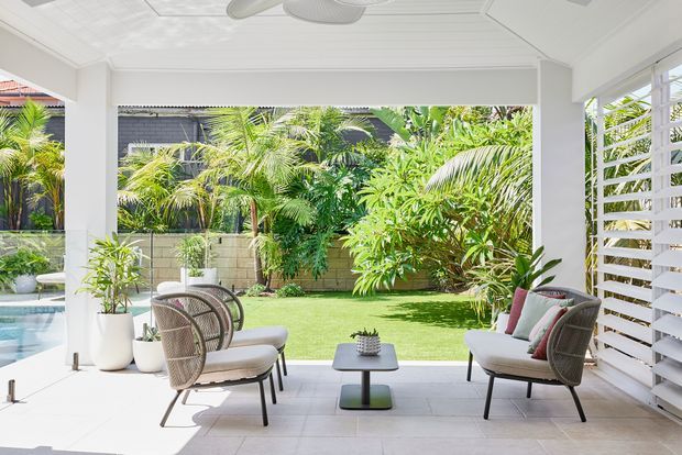 Bright covered outdoor sitting area opening onto a lush landscaped garden and swimming pool. The space features white structural framing, a tiled floor, louvred screening on the right, woven lounge chairs with cushions, and a small central table. The setting has a relaxed tropical-resort feel with strong indoor–outdoor connection.