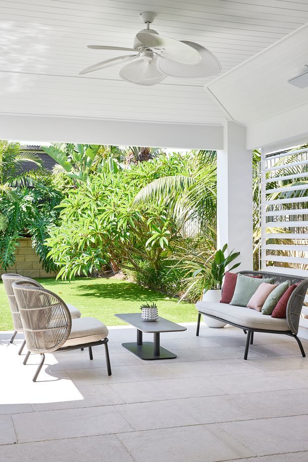 Bright covered outdoor living area with a white-lined pitched ceiling, sculptural ceiling fan, tiled floor, louvred side screening, and contemporary outdoor seating arranged around a low coffee table. The open space overlooks a lush tropical garden and lawn.