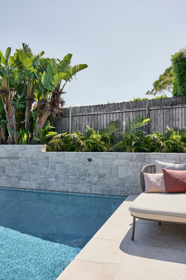 A bright landscaped pool area with a blue swimming pool bordered by pale stone paving. A raised grey stone retaining wall and timber privacy fence frame tropical planting, while a cushioned outdoor lounge chair sits beside the pool.