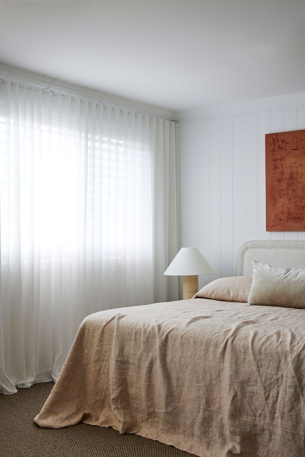 Bright, softly styled bedroom featuring a partially visible upholstered bed with textured beige linen bedding, sheer full-height curtains over a large window, white vertical wall panelling, a woven bedside lamp, and a warm rust-toned artwork. The room has a calm, neutral palette and abundant diffused natural light.