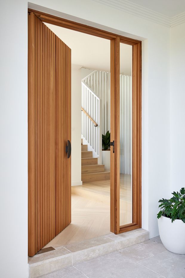 A bright residential entrance viewed from outside, featuring a tall timber pivot door with vertical slatted detailing, a matching timber-framed glazed sidelight, and a pale stone threshold. Beyond the doorway is a light-filled hallway with timber flooring, a white staircase and balustrade, and indoor planting. White walls with decorative ceiling cornices create a refined, contemporary-traditional entry.