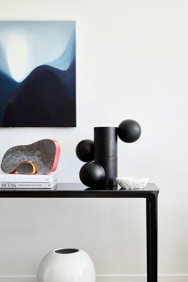A minimalist interior vignette featuring a slim black console table against a white wall. The console displays a black sculptural object with spherical forms, a textured abstract artwork in deep blue and pale tones, stacked design books, a small decorative bowl, and a white ceramic vessel below. The composition is monochromatic with sculptural contemporary decor and clean lines.