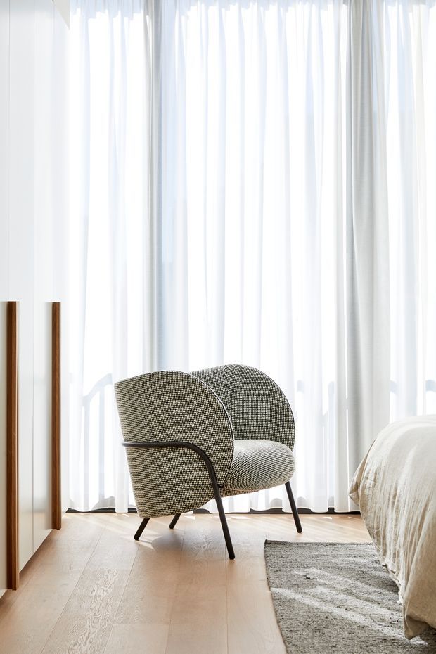 Bright minimalist bedroom corner featuring a sculptural upholstered armchair with dark slender legs, sheer full-height white curtains, pale timber flooring, a partially visible upholstered bed, and a textured grey rug. White built-in cabinetry with vertical timber handles occupies the left side. The space has abundant diffused natural light and a calm, refined atmosphere.