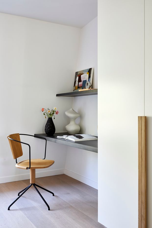 A compact, bright home office nook with a timber-and-black metal swivel chair, pale timber flooring, white walls, floating dark shelves, decorative objects, and a tall white built-in cabinet with a vertical timber handle. The space has a clean, refined, minimalist appearance with subtle mid-century-inspired furniture.