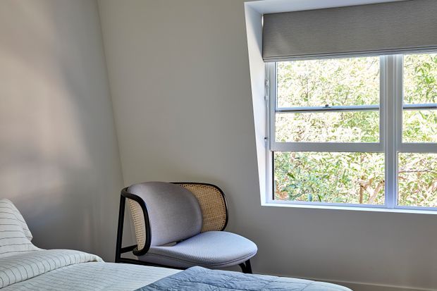 A softly lit bedroom corner with a partially visible bed in the foreground and a modern upholstered lounge chair positioned beside a large white-framed window. The window has a roller blind and overlooks leafy greenery, while the room features plain light neutral walls and a calm, understated interior.