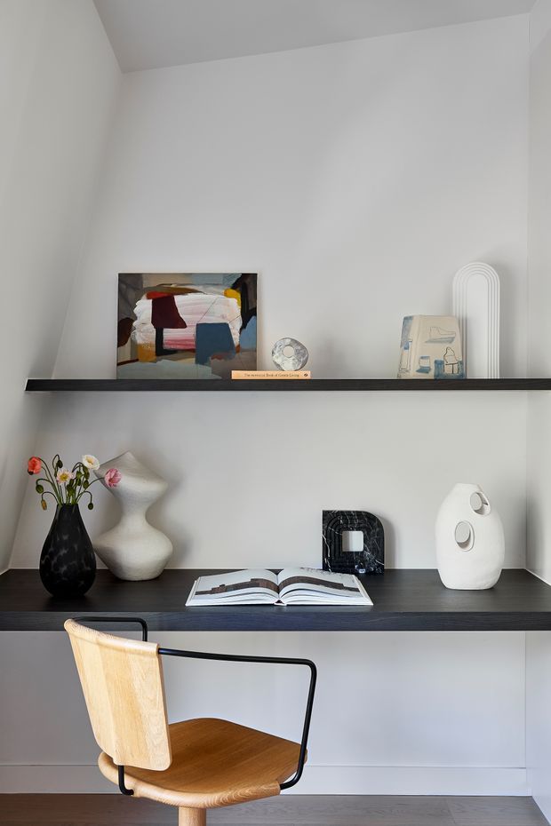A minimalist built-in study nook with white walls and dark floating shelves. The shelves display artwork, books, sculptural ceramics, and a vase with flowers, while a simple wooden and black metal chair sits at the desk below. The space is bright, clean, and restrained, with a high angled ceiling and pale timber flooring.