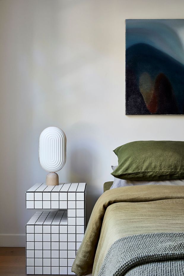 A close-up bedroom interior with a neatly made bed in muted olive green and beige bedding. Beside it is a distinctive white grid-tiled bedside table with an open lower shelf, topped by a sculptural white lamp. A dark abstract artwork hangs above the bed area against a pale neutral wall, with warm timber flooring visible at the bottom.