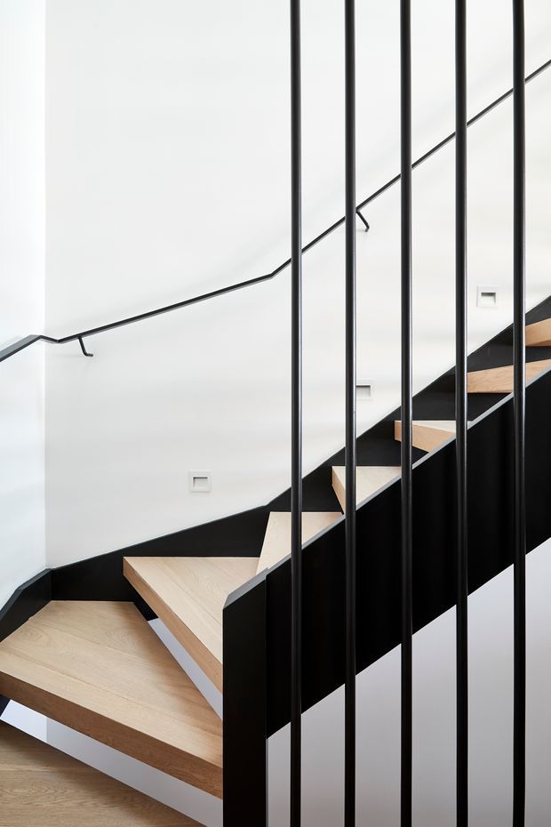 A minimalist interior stairway with pale timber treads, dark painted stringers and side panels, white walls, and slender black vertical balustrade bars. A simple black handrail follows the stair and wall, creating a strong graphic contrast.