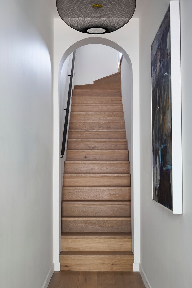 A narrow, refined stairway viewed from a hallway, featuring warm oak timber treads and risers, white painted walls and trim, a black handrail, and a rounded arched opening. A large woven black pendant light hangs overhead, with abstract artwork mounted on the right wall.
