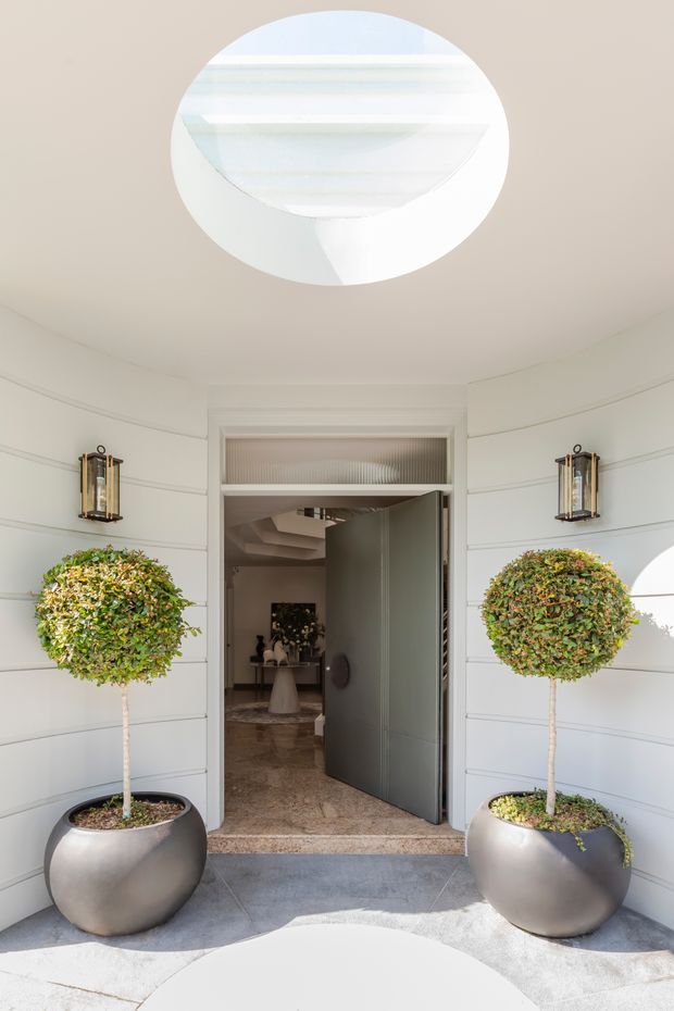 Bright, symmetrical residential entryway with white horizontal-panel cladding, a large circular skylight, matching spherical topiary trees in dark rounded planters, decorative wall lanterns, and a wide dark entry door opening into a refined interior. The interior beyond includes a sculptural staircase and console styling.