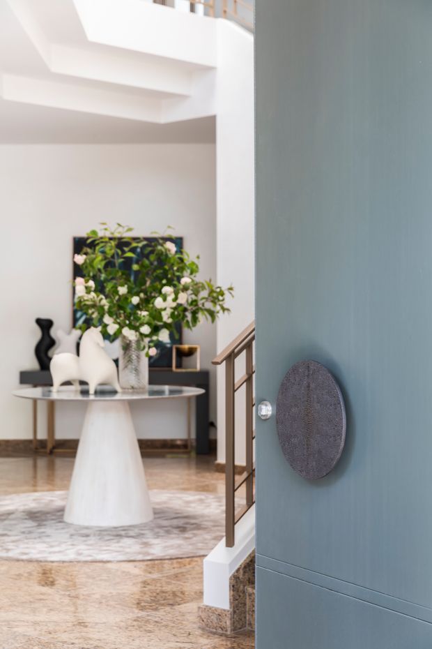 A bright, refined residential entryway viewed through a partially open blue-grey door. The foyer features a sculptural white pedestal table, decorative objects, a large floral arrangement, a console, framed artwork, a patterned rug, polished stone flooring, and a staircase with a timber handrail and dark metal balustrade. A high-level landing and open void contribute to the airy spatial quality.
