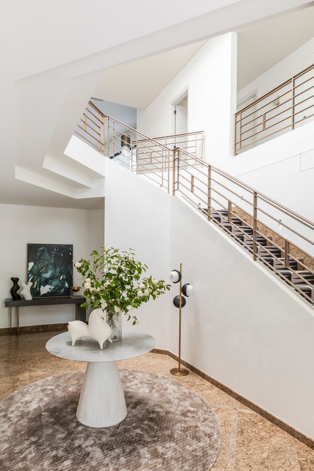 Bright, refined residential entry hall with a dramatic multi-level stairway and open void. White walls and ceilings are paired with slim brass-toned geometric railings, polished stone flooring, a round sculptural table, contemporary artwork, decorative objects, and a tall floor lamp. A large softly patterned rug and indoor plant add warmth to the otherwise minimalist interior.