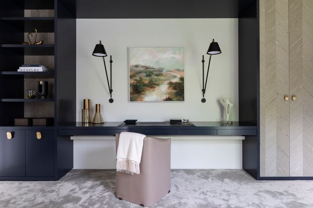 A refined home office or study with a built-in dark joinery wall, integrated desk, open shelving, concealed storage, and pale timber cabinetry. Two black wall-mounted lamps flank an abstract landscape artwork, while a soft upholstered chair and light patterned rug add warmth and contrast. The room has a restrained, contemporary palette of charcoal, soft white, taupe, and pale wood.